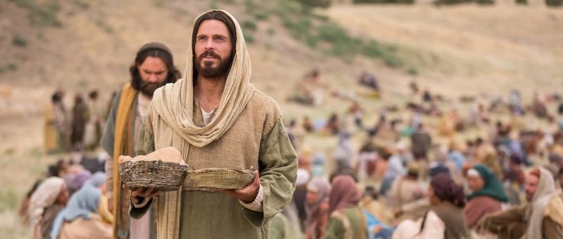 A still image from the Bible Video Series video of Jesus feeding the 5,000. Jesus stands with a basket of five loaves and two fishes, giving thanks to God.